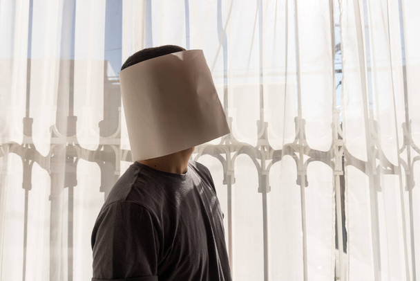 Młody Latynos w białej papierowej masce zakrywającej całą twarz. Czysta kartka. Stoi przy oknie w kostiumie. Słoneczny dzień w Bogocie, Kolumbia. - Zdjęcie, obraz