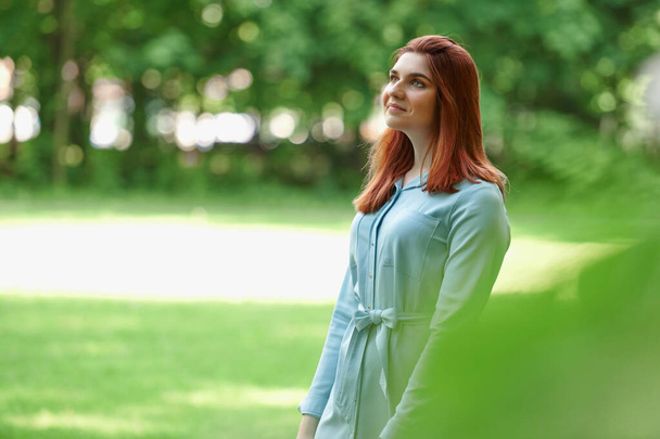 portrait of a beautiful girl with red hair and a blue dress on a green natural background in summer. The girl stands in the branches of a green tree, looks away and smiles - Фото, изображение