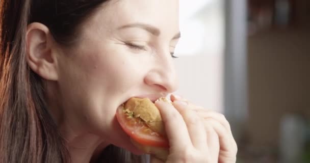 mooie vrouw eten cheeseburger met genot en verwennen met de smaak binnen met zon schijnt een close-up schot op rode camera - Video