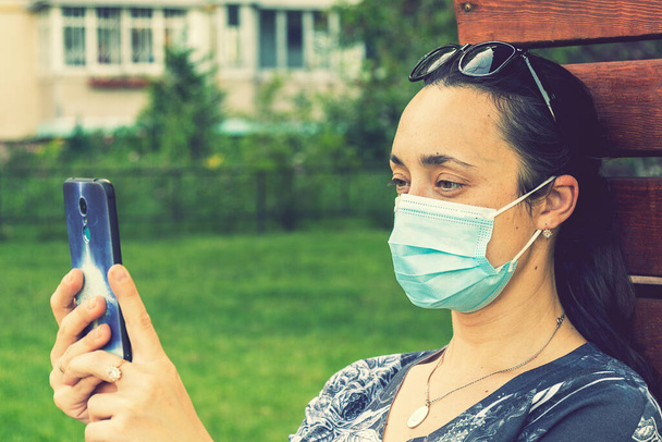 Koruyucu maskeli genç bir kadın akıllı telefon tutuyor. Green Park 'ta bankta oturan cep telefonlu bir kadın. Koronavirüs kısıtlamalarının gevşetilmesi ve kendini izole etmenin ilk aşaması. ses tonlu - Fotoğraf, Görsel