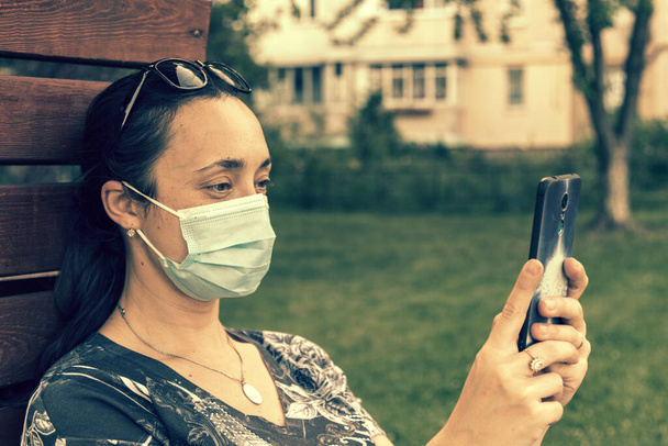 Ritratto di una ragazza con maschera medica al parco con smartphone durante la quarantena. Una giovane ragazza con una maschera medica siede sulla panchina del parco e usa lo smartphone. tonica. - Foto, immagini