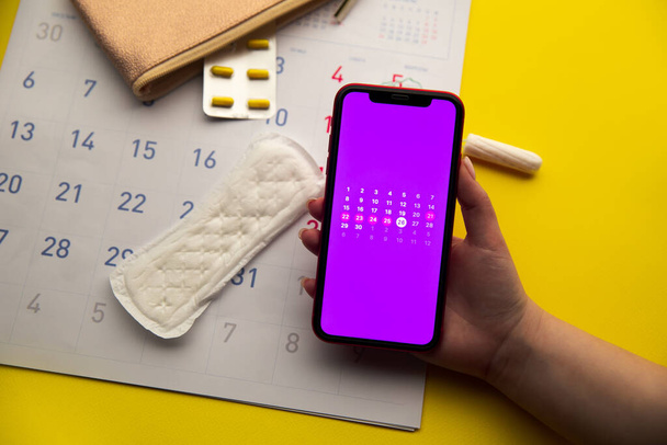 Menstrual cycle concept. Calendar for the month with marks and a mobile application on the smartphone screen. Contraceptive and sanitary pads on the background. - Photo, Image