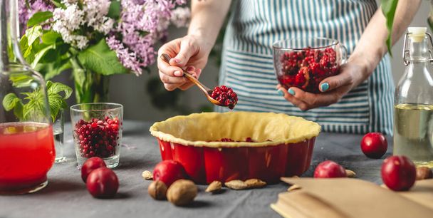 Μαγειρεύω σπιτική πίτα βατόμουρο. Στο ταψί με τη ζύμη βάζουμε ένα κουτάλι με φρέσκα cranberries. Τραπέζι με φωτεινά συστατικά σε ανοιξιάτικο στυλ με λουλούδια - Φωτογραφία, εικόνα