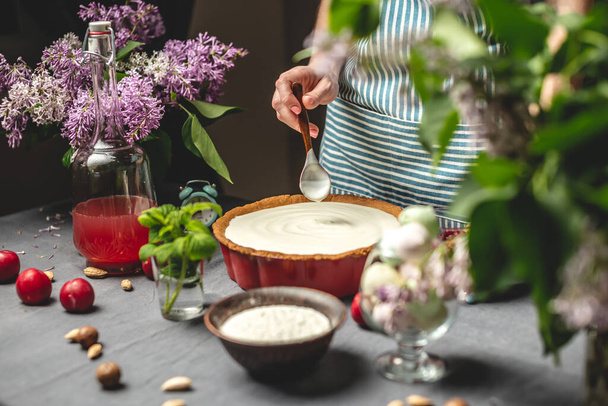 Cooking homemade cranberry pie. A female pastry chef is adding of sour cream with spoon to a baked tart with lingonberries. Table with bright ingredients in spring style with flowers - Photo, Image