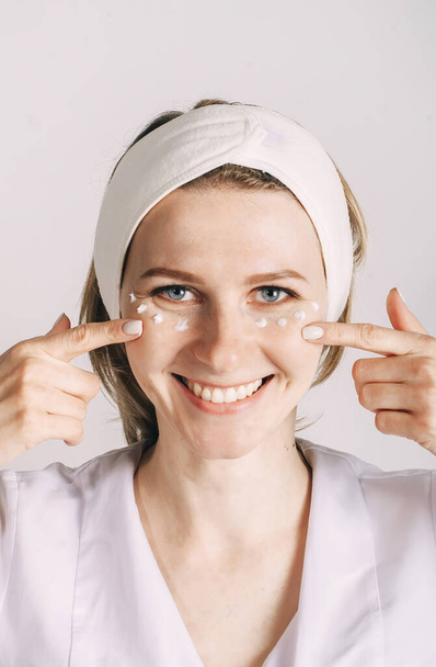 Smiling young woman apply moisturizing cosmetic cream on face look at camera with towel on head. Girl care of healthy clean skin use spa treatment lotion. Closeup portrait - Photo, Image