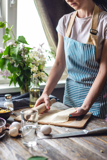 Nainen levittää tuoretta taikinaa pastan valmistukseen viihtyisässä ilmapiirissä. Käsite ruoanlaitto maukasta ruokaa kotona - Valokuva, kuva