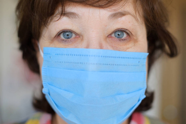 An aged woman in a protective medical mask is looking directly at the camera. Pensioner in self-isolation for the period of quarantine at the height of the virus - Фото, изображение
