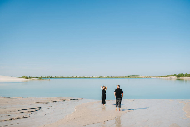 jong paar een jongen met een meisje in zwarte kleren zijn lopen op het witte zand aan de rand van blauw water - Foto, afbeelding