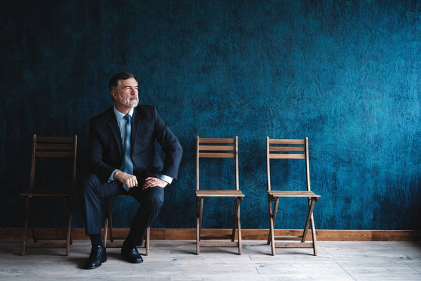 Waiting for interview. Confident mature businessman sitting on chair against dark blue background. - Foto, afbeelding
