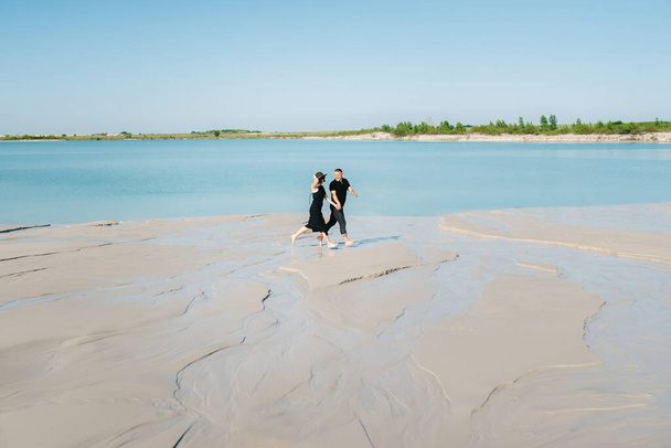 young couple a guy with a girl in black clothes are walking on the white sand at the edge of blue water - Photo, Image