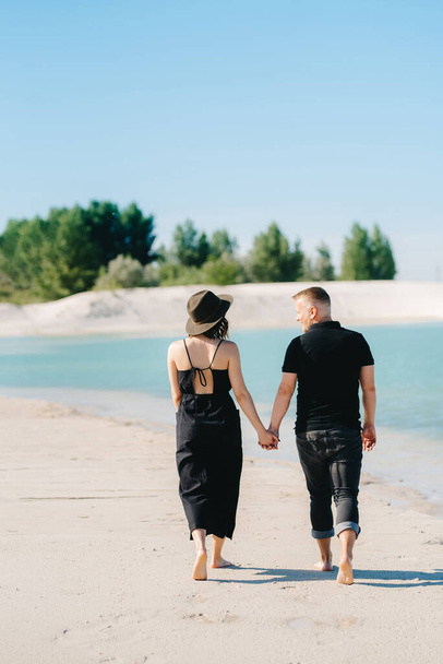 黒い服を着た女の子を持つ若いカップルが青い水の端の白い砂の上を歩いています - 写真・画像