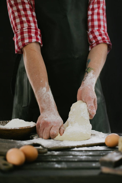 Mooie en sterke mannenhanden kneden het deeg waaruit ze dan brood, pasta of pizza zullen maken. Een wolk bloem vliegt rond als stof. Naast het kippenei. De achtergrond is donker - Foto, afbeelding