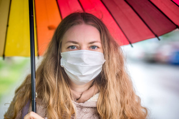 Caucasian girl in a protective mask walks under an umbrella on an empty street in spring rain. Safety and social distance during a coronavirus pandemic. New normal, the implications of quarantine. - Foto, imagen