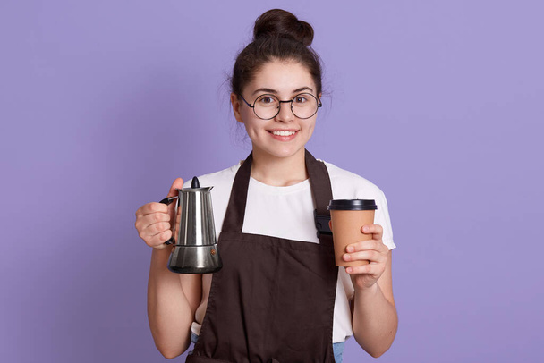 Smiling waitress in white t shirt and brown apron holding pot and take away cup in hands, looks at camera with happy facial expression, woman with positive expression. - Photo, image