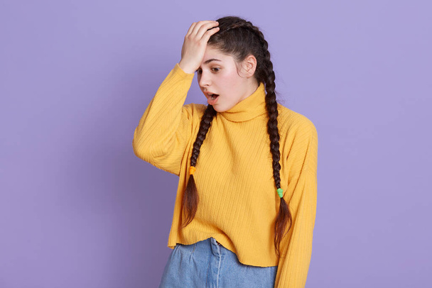 Girl with long dark hair and pigtails standing with opened mouth, keeping hand on forehead, forget something important, woman standing against lilac wall. - Foto, Bild