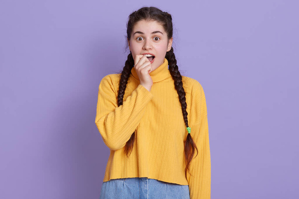 Excited woman bites her fist and looks at camera with dropped eyes, lady looks astonished, wearing yellow sweater, posing isolated over lilac background. - Fotoğraf, Görsel