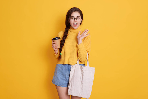 Young stylish woman wearing shirt and short, lady holding bag and take away coffee, female has long dark hair, posing with opened mouth, girl enjoying hot beverage during shopping. - Foto, imagen