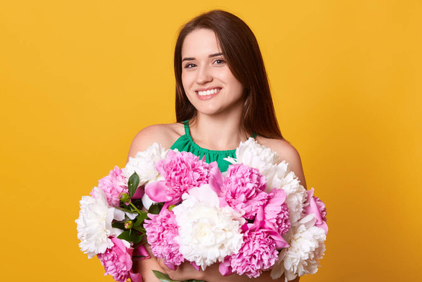 Smiling brunette woman standing in green attire against yellow background, holding beautiful flowers, girl looking at camera with happy facial expression. - Photo, image