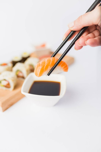 Close up of woman holding chopsticks with salmon sushi roll and bowl with soy sauce on table, portion of traditional japanese meal in the background. - 写真・画像