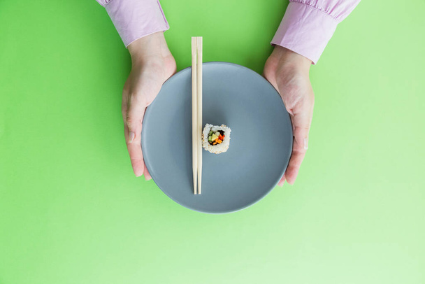 Płaskie układanie żeńskich dłoni trzymając płytę z pałeczkami i jedną rolkę sushi izolowane na zielonym tle. - Zdjęcie, obraz
