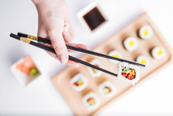 Close up of female hand holding sushi roll with chopsticks above sushi set portion on table. - 写真・画像