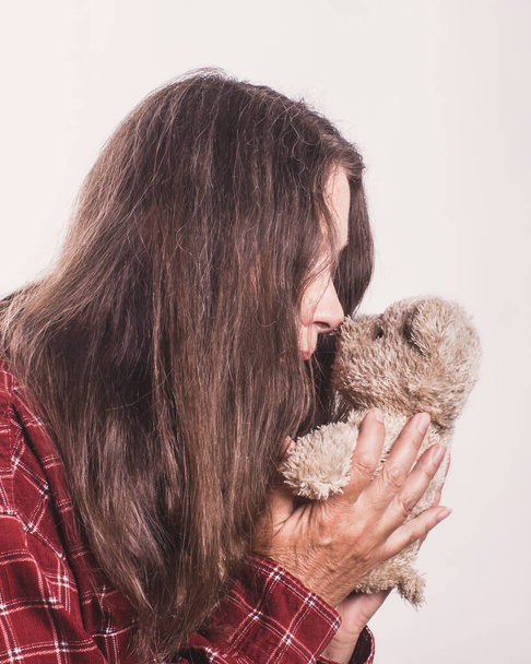 Μια ηλικιωμένη γυναίκα με μακριά γκρίζα μαλλιά δείχνει αγάπη και αφοσίωση στο αρκουδάκι της. Πορτρέτο στο προφίλ, κοντινό πλάνο. - Φωτογραφία, εικόνα