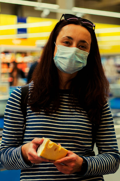 donna in maschera medica protettiva in un supermercato sceglie il formaggio. il concetto di pandemia e covid-19. Mano dell'acquirente con un pezzo di formaggio nel negozio. tonica. foto verticale. - Foto, immagini