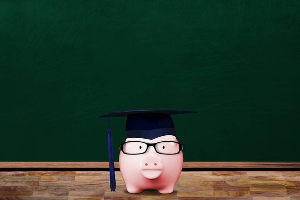 Saving for education with pink piggy bank wearing graduate hat and eyeglasses in school classroom with copy space on chalkboard. Concept of education investment, tuition cost, college loan, etc. - Photo, Image