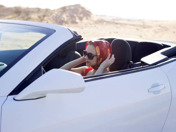 Retrato de bela glamour jovem mulher sentar-se no descapotável cabriolet esporte carro branco auto e corrigir seu cachecol vermelho seda no espelho, usando óculos de sol, cabeça de cobertura com cachecol de seda, roupa de luxo moda vermelha
  - Foto, Imagem