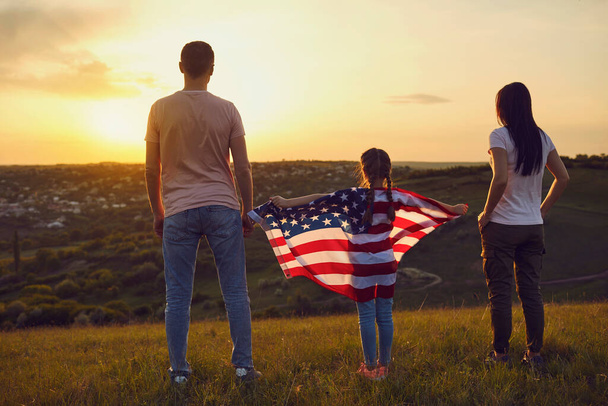 Happy family with usa flag on nature in the evening at sunset. Back view. Mother father and daughter celebrate fun Independence Day of America. - Фото, зображення