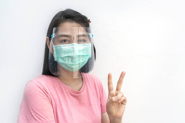 Απομονωμένο πορτραίτο Ασιάτισσας που φοράει ιατρική μάσκα προσώπου και ασπίδα προσώπου σε λευκό φόντο, δείχνει χειρονομία δύο δάχτυλα, μαύρα κοντά μαλλιά και φοράει μπλουζάκι, κοίτα κάμερα, Περικοπή διαδρομής στο αρχείο. - Φωτογραφία, εικόνα