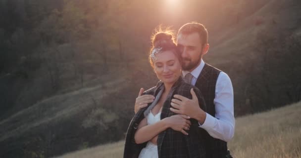 Close-up shot of cute married millennial couple hugging in a deserted spikelet field on warm sunny day. Carring groom wraps his beautiful bride in elegant white wedding dress with jacket. Rest outside - Footage, Video