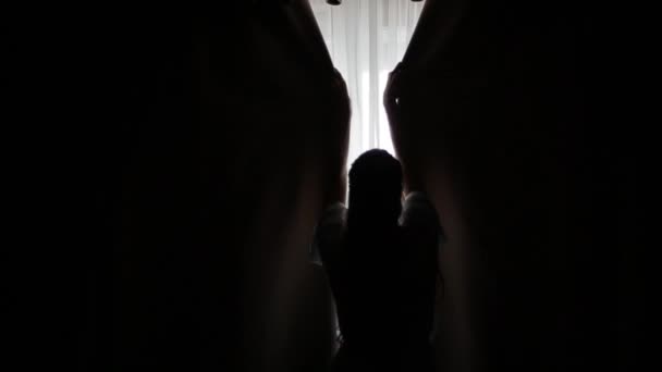 Zadní záběr kavkazské nevěsty v průhledných tyrkysových županech s dlouhými vlasy, které rozhrnou záclony u okna a ráno osvětlí tmavý pokoj. Dívka odhrnuje černé závěsy. - Záběry, video