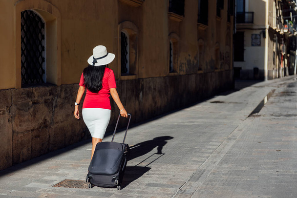 Latin woman are hat walking away from behind dragging a suitcase suitcase down a street in Europe. travel concept - Фото, изображение