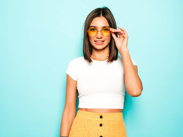 Young beautiful woman looking at camera.Trendy girl in casual summer white T-shirt and yellow skirt in round sunglasses. Positive female shows facial emotions. Funny model isolated on blue - Фото, зображення