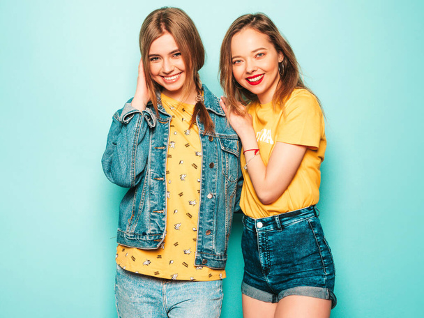 Две молодые красивые улыбающиеся хипстерки в модных летних желтых футболках и джинсовой куртке. Сексуальные беззаботные женщины позируют у голубой стены. Традиционные и позитивные модели развлечения - Фото, изображение
