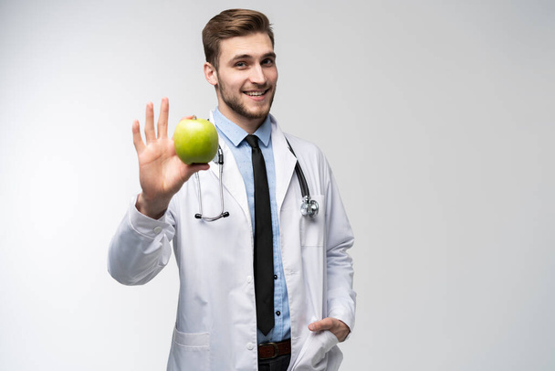 Portrait of handsome doctor in white coat looking at camera and smiling while holding an apple, isolated on white. - Foto, imagen