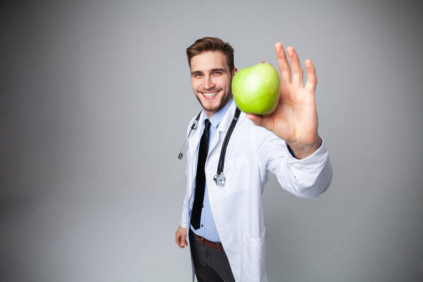 Portrait of handsome doctor in white coat looking at camera and smiling while holding an apple, isolated on white. - Photo, Image