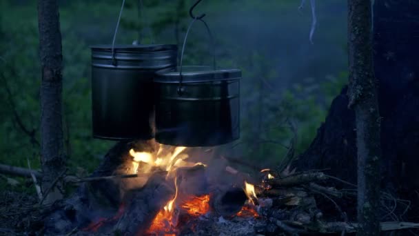 Vacsorát főzni a tábortűznél, az éjszakai ködös erdőben, túrán. Fekete turisztikai fazekak lógnak égő tábortűz felett sötét erdőben. - Felvétel, videó