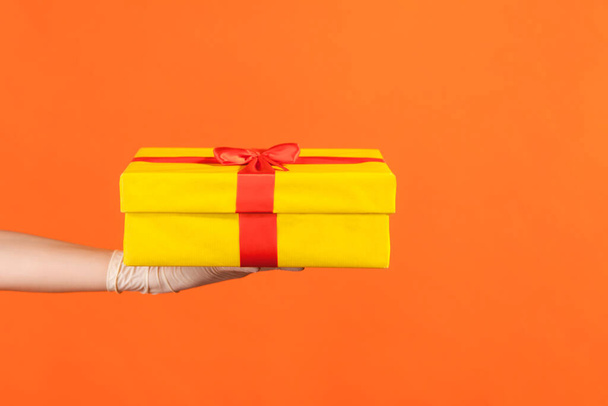 Profile side view closeup of human hand in white surgical gloves holding yellow gift box. indoor, studio shot, isolated on orange background. - Photo, image