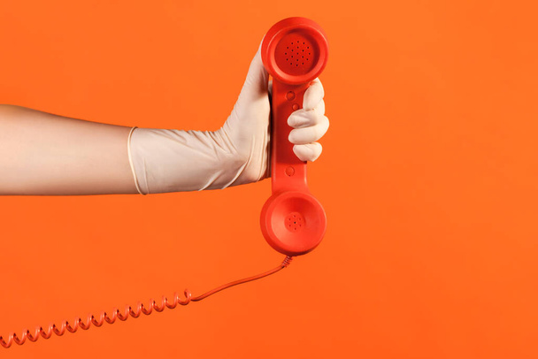 Profile side view closeup of human hand in white surgical gloves holding and showing red call telephone handset receiver. indoor, studio shot, isolated on orange background. - Fotó, kép