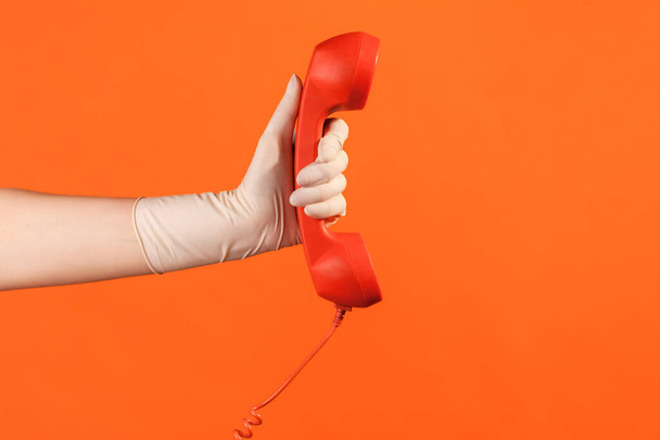 Profile side view closeup of human hand in white surgical gloves holding and showing red call telephone handset receiver. indoor, studio shot, isolated on orange background. - Fotoğraf, Görsel