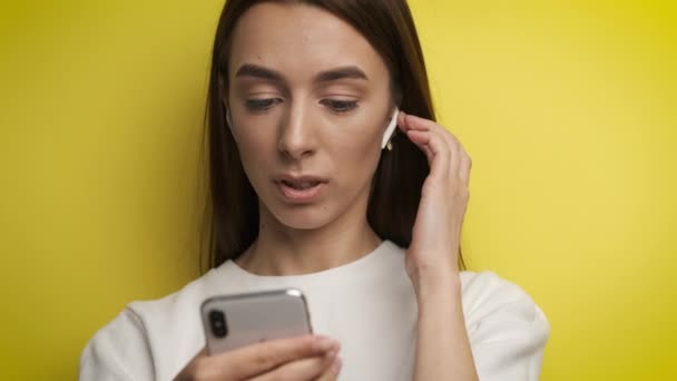 Beautiful 25 year old woman on a yellow background holds a phone in her hands and communicates by video call through headphones - Imágenes, Vídeo