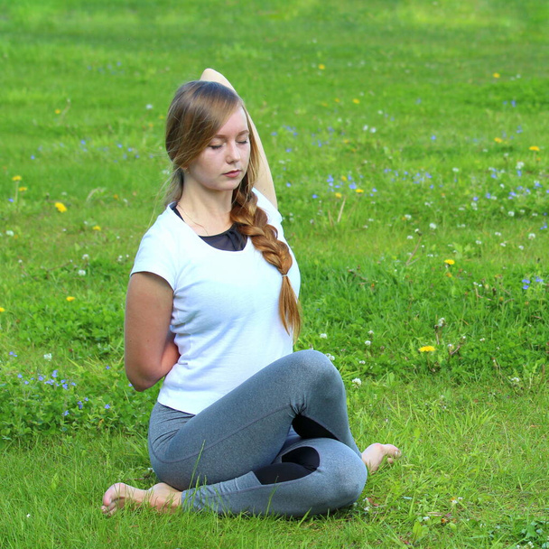 Young woman of European appearance does yoga in summer nature. Woman sitting in cow face pose, gomukhasana. High quality photo for web and print with empty space for text and design. - Foto, imagen