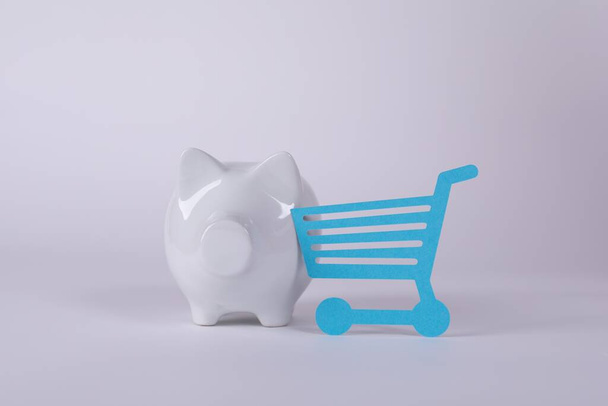A closeup shot of a blue shopping cart paper cut-out near a white piggy bank on the table - Φωτογραφία, εικόνα