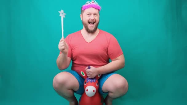 Een jonge man met een baard in een roze T-shirt met een diadeem op zijn hoofd op een eenhoorn met een toverstaf in zijn hand. Een grappige tovenaar grap te maken en een wens te vervullen. - Video