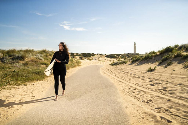 A shallow focus shot of an attractive female carrying a surfboard while walking in the middle of the road in Spain - Фото, зображення