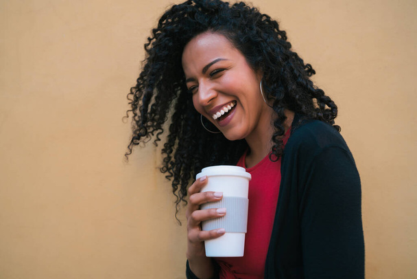 Portrait of young afro american woman looking confident and posing while holding a cup of coffee against yellow background. - Fotó, kép