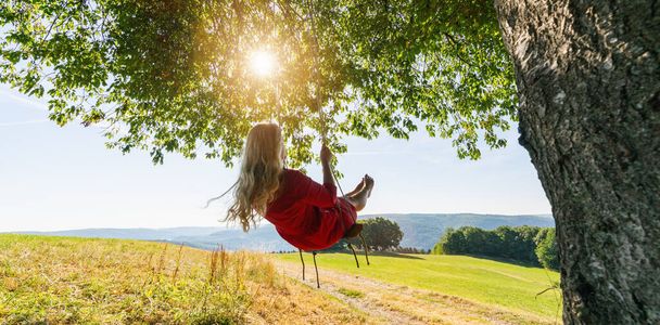 Nerozpoznatelná dívka v červených šatech houpající se na houpačce na stromě za klidného večera. Dáma sedí na dřevěném houpačce a dívá se na zlatý západ slunce. Mladá žena houpající se při východu slunce - Fotografie, Obrázek
