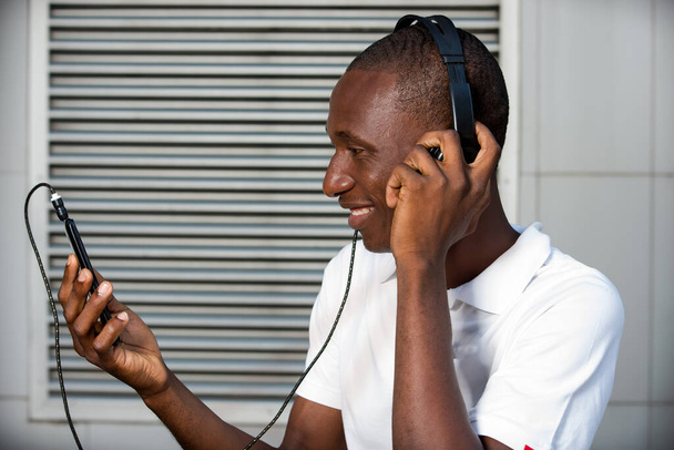щасливий молодий чоловік у білій футболці слухає музику з гарнітурою мобільного телефону і співає на відкритому повітрі. Студент зі смартфоном та навушниками
 - Фото, зображення